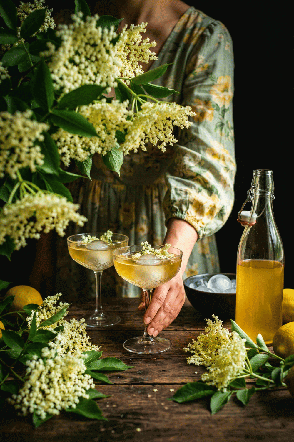 A world-class luxury overhead dark moody food photograph of elderflower cordial presentation. Shot at exactly 90 degrees overhead. Centered on a tarnished antique silver tray with soft patina marks, its surface lined with rumpled jet-black velvet fabric that absorbs light into deep shadow. Inside the tray, three vintage clear glass swing-top bottles with wire bales and porcelain stoppers rest at lazy angles&mdash;one large bottle reclining on its side, two smaller apothecary bottles nestled close. All filled with brilliant bright golden-yellow translucent elderflower cordial (Sambucus nigra) that glows like molten sunshine against the dark velvet, the liquid intensely saturated and luminous. No labels on bottles. Abundant fresh elderflower umbels with creamy-white lacy florets and bright green stems arranged in wild profusion&mdash;clusters spilling from the tray onto the surrounding dark weathered wood surface, some blooms resting directly on the black velvet, others framing the bottles naturally. A few delicate white cow parsley (Anthriscus sylvestris) stems and soft green fern fronds mixed throughout. Loose individual florets dust the velvet like pale snowflakes. Single dramatic natural window light streams from upper left, heavily flagged to create authentic chiaroscuro&mdash;the beam strikes the vibrant golden cordial making it glow brilliantly against the darkness while the surrounding velvet and wood fall into deep velvety black shadow. Rich contrast between the bright luminous liquid and the enveloping darkness. The black velvet catches only subtle rim light. No window visible. The composition feels abundant, wild, and luxuriously mysterious&mdash;like discovering a secret harvest in a dim garden room. Shallow depth of field with sharp focus on the glowing golden liquid and bottle glass, brilliant warm golden-yellow highlights of the cordial contrasting against deep charcoal blacks and tarnished silver, atmospheric storytelling composition, professional editorial food photography, 8k resolution. Old Fashioned Elderflower Cordial Recipe